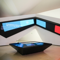 Área Amanhãs da Exposição Principal, com paredes em formato origami, coberta por telas em ângulos e, ao centro, uma mesa interativa