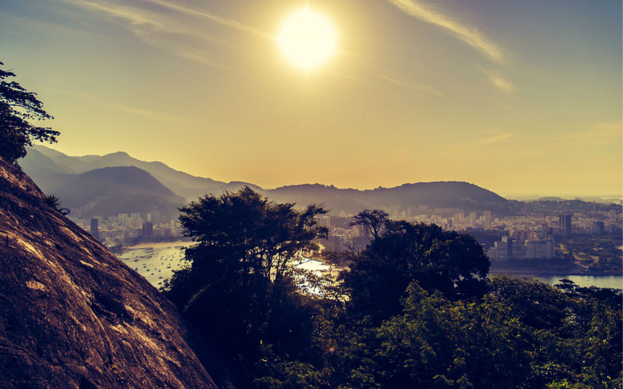 Final de tarde no Rio de Janeiro visto de cima de um morro. A perspectiva da foto mostra a floresta na frente do mar da cidade.