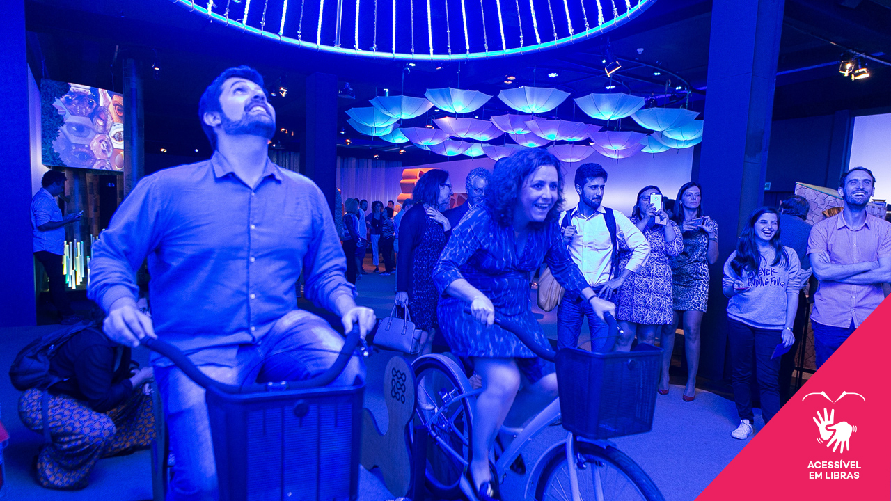 Duas pessoas na área de colaboração da exposição Inovanças, andando de bicicleta para fazerem um lustre com luz azul acender