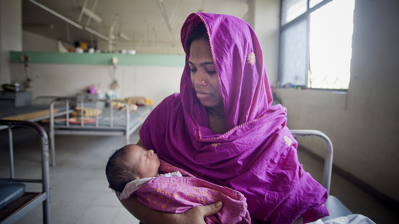 Mulher com trajes tradicionais segurando um bebê recém-nascido. Ao fundo, o que parece ser um hospital sem luxos.