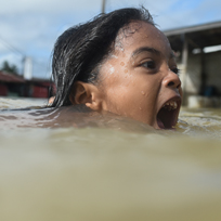 Menina nadando em água aparentemente suja e com a boa aberta pedindo ajuda