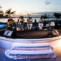 Foto feita na lateral do museu, com banda de música tocando em palco circular branco e, ao fundo, a Baía de Guanabara no pôr do sol.