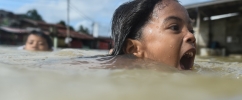 Menina nadando em água aparentemente suja e com a boa aberta pedindo ajuda